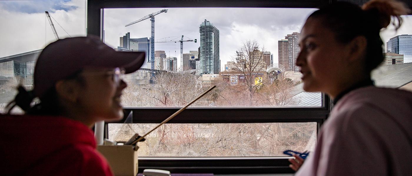 Two students talking in CDES Suite, with view of Denver skyline seen through a window in the background