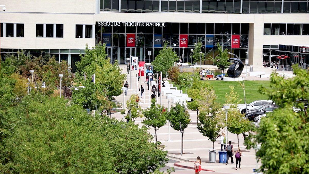 An aerial view of the MSU Denver campus, focusing on the Jordan Student Success Building. The entrance is busy with students walking along the pathways lined with trees. The building has large glass windows and 'Jordan Student Success' is written above the entrance. A sculpture is visible on the lawn, and a coffee shop with outdoor seating is on the left.